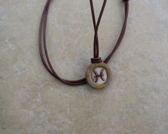 May include: A brown leather cord necklace with a round ceramic pendant featuring the Pisces zodiac symbol in white.