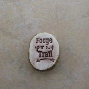 May include: A white stone with a brown inscription that reads "Forge your own Trail" with an arrow pointing to the right.