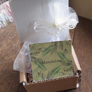 May include: Open cardboard box with a white interior and a white tulle bag tied with a ribbon. A card with the text "Mareziedotes" and a green leaf pattern is inside the box.