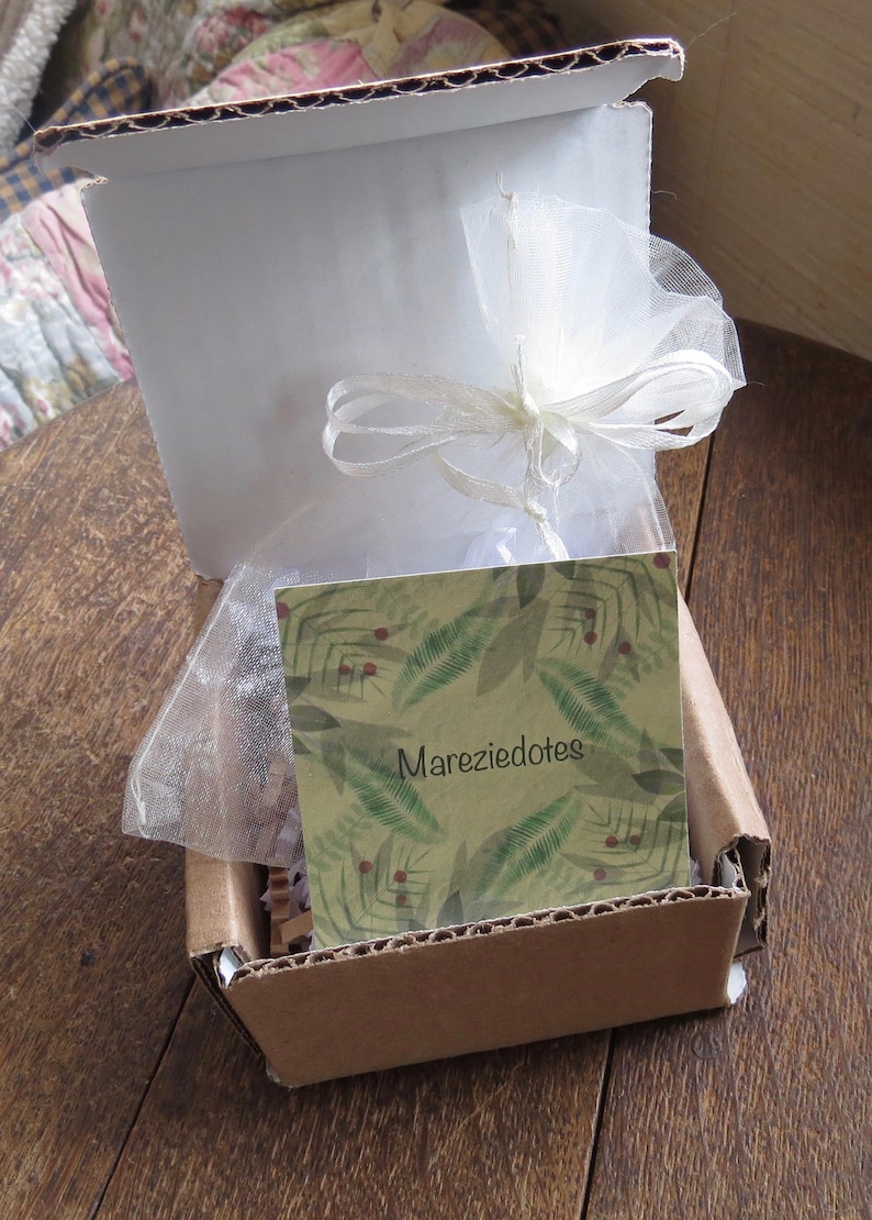 May include: Open cardboard box with a white interior and a white tulle bag tied with a ribbon. A card with the text "Mareziedotes" and a green leaf pattern is inside the box.