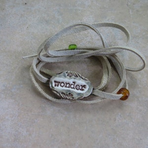 May include: A handmade leather wrap bracelet with a ceramic charm. The charm is oval-shaped with the word "wonder" in brown lettering. The bracelet has a light tan color and features a green and a brown bead.
