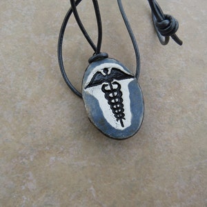 May include: A black leather cord necklace with a blue and white ceramic pendant featuring a medical symbol, the Rod of Asclepius, with a snake wrapped around it and wings on top.
