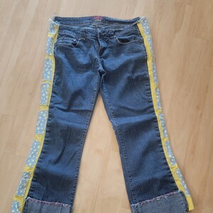 May include: Blue denim jeans with yellow and blue star-shaped embroidery on the sides. The jeans have a wide leg and are cuffed at the bottom.