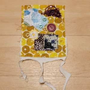 Może przedstawiać: Żółty kwadratowy kawałek materiału z białą obwódką i różnymi kawałkami materiału przyszytymi na wierzchu. Kawałki obejmują materiał w niebiesko-białe wzory, materiał kwiatowy i materiał czarno-biały. Są też dwa guziki, jeden niebieski i jeden czerwony, przyszyte do materiału.