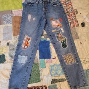 Puede incluir: Vaqueros azules de mezclilla con múltiples parches de diferentes telas y colores cosidos. Los vaqueros tienen áreas desgastadas y bordes deshilachados.