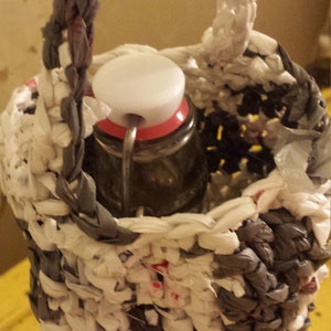 May include: A crocheted bottle holder made from recycled plastic bags. The holder is white and grey with a black bottle inside.