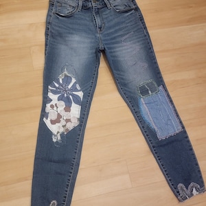 May include: Blue denim jeans with decorative patches and floral designs. The jeans have a high waist and a distressed hemline. Patches include a floral design and a light blue denim patch.