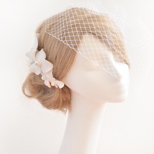 May include: White bridal hair comb with delicate flower accents and pearl embellishments. The comb is worn in a blonde updo with a white birdcage veil.