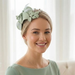 Puede incluir: Una mujer con un vestido verde claro y un tocado floral a juego. El tocado presenta flores de tela y detalles de perlas. La mujer sonríe y el fondo es de color neutro.