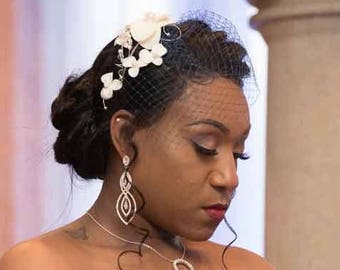 Op de afbeelding: Een witte strapless trouwjurk met ingewikkeld kralenwerk en borduursels. Het haar is in een opgestoken kapsel met een witte bloemenhaaraccessoire en een korte sluier. Een zilveren ketting met een ronde hanger en lange oorbellen.