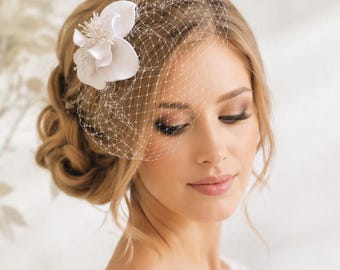 Op de afbeelding: Een wit birdcage sluier haaraccessoire met een delicaat netpatroon en een enkele grote witte stoffen bloem met gedetailleerde meeldraden, ontworpen om als decoratief hoofddeksel voor formele gelegenheden te dragen.