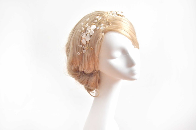 May include: A hair vine accessory featuring gold-toned wire branches adorned with small white flower petals, clear crystal beads, and tiny gold accents. The piece is arranged in a delicate, branching pattern designed to be worn along the side of the hair.