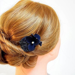 May include: A woman's hair is styled in a low bun with a navy blue flower hair clip. The flower has a pearl in the center.