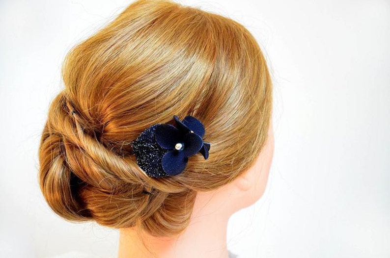 May include: A woman's hair is styled in a low bun with a navy blue flower hair clip. The flower has a pearl in the center.