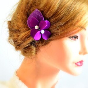 May include: A purple flower hair clip with pearl accents. The flower is made of fabric and has a sparkly texture.