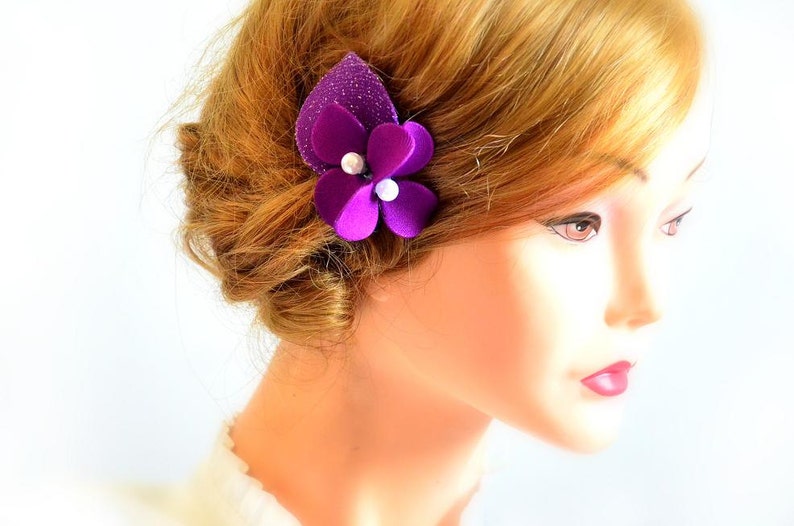 May include: A purple flower hair clip with pearl accents. The flower is made of fabric and has a sparkly texture.