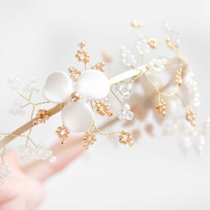 May include: A gold-toned metal headband featuring a central white fabric flower with a small gold bead at its center. The band is adorned with delicate gold wire branches decorated with clusters of small clear and metallic gold-colored beads.