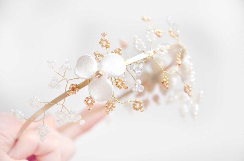 May include: A gold-toned metal headband featuring a central white fabric flower with a small gold bead at its center. The band is adorned with delicate gold wire branches decorated with clusters of small clear and metallic gold-colored beads.