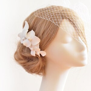 May include: White floral hair comb with pearl accents. The comb is worn in a blonde updo with a white veil.