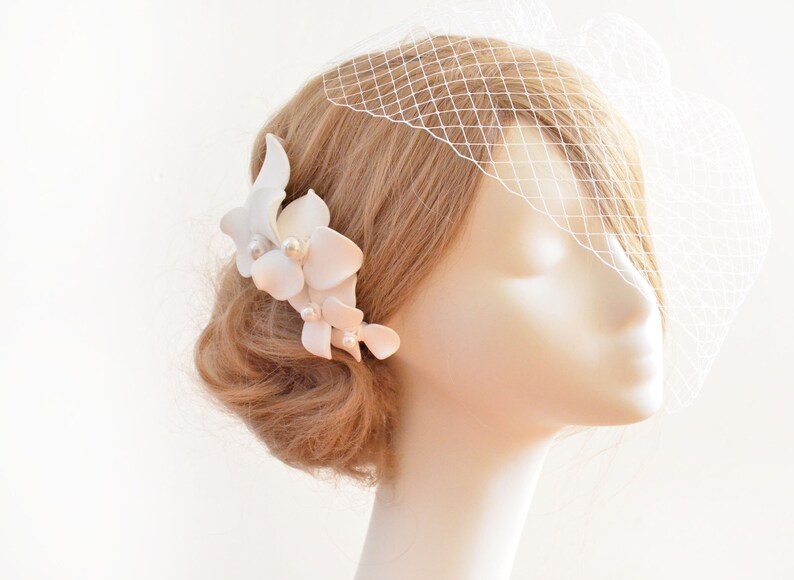 May include: White floral hair comb with pearl accents. The comb is worn in a blonde updo with a white veil.