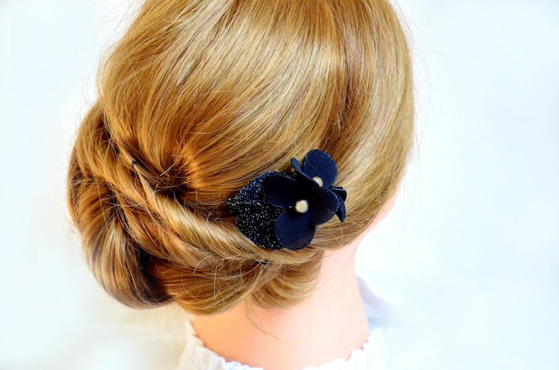 May include: A black fabric flower hair clip with a sparkly center. The clip is worn in a woman's blonde hair.