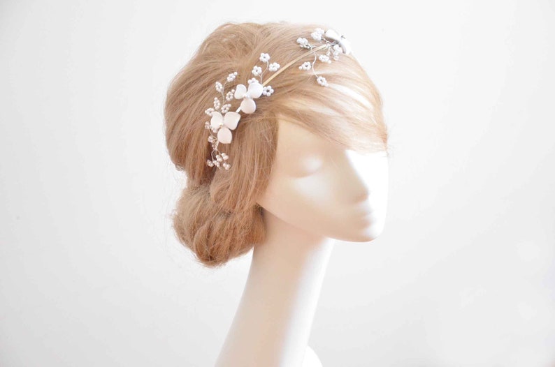 May include: A gold-toned metal headband featuring delicate wire branches adorned with small white floral accents and tiny pearl-like beads. The flexible wire structure allows for intricate positioning across the hair, showcasing a botanical design.