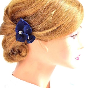 May include: A blue velvet flower hair clip with a rhinestone center. The clip is worn in a woman's hair.