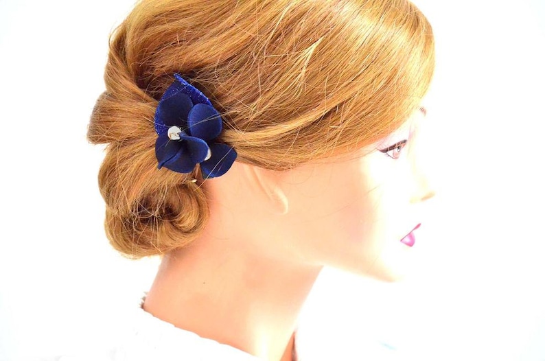 May include: A blue velvet flower hair clip with a rhinestone center. The clip is worn in a woman's hair.