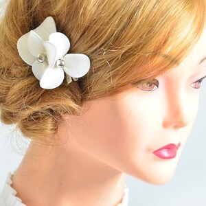 May include: White flower hair clip with pearl accents. The clip is made of fabric and is attached to a silver metal clip.
