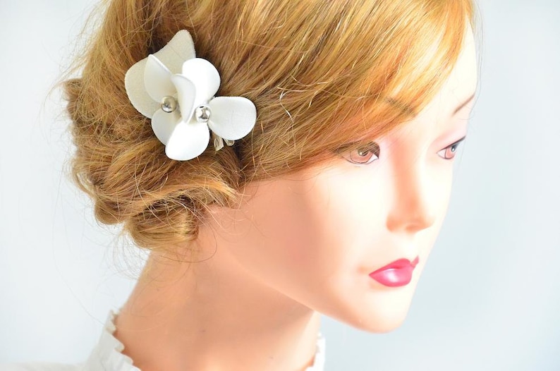 May include: White flower hair clip with pearl accents. The clip is made of fabric and is attached to a silver metal clip.