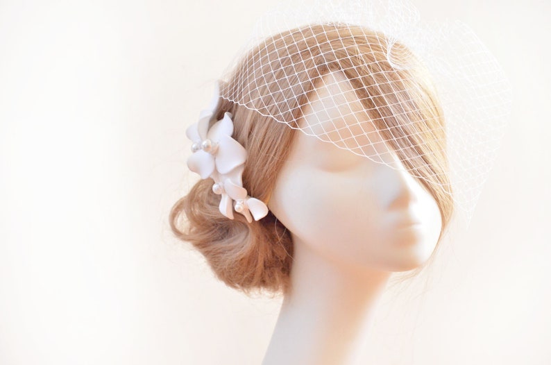 May include: A white bridal veil with a delicate floral hairpiece. The veil is made of a fine mesh fabric and is attached to a comb. The hairpiece features white flowers and pearls.