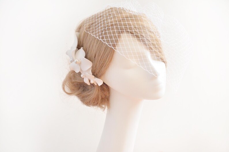 May include: White bridal hair comb with delicate flower accents and pearl embellishments. The comb is worn in a blonde updo with a white birdcage veil.