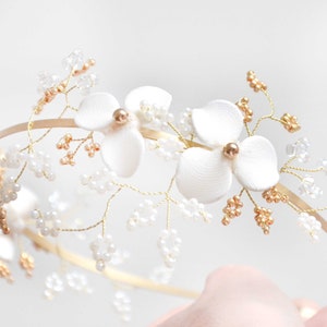 May include: A gold-toned metal headband featuring two white fabric flowers with gold centers, accented by delicate gold wire branches adorned with small clear beads and tiny gold-colored beads, creating a botanical-inspired hair accessory.