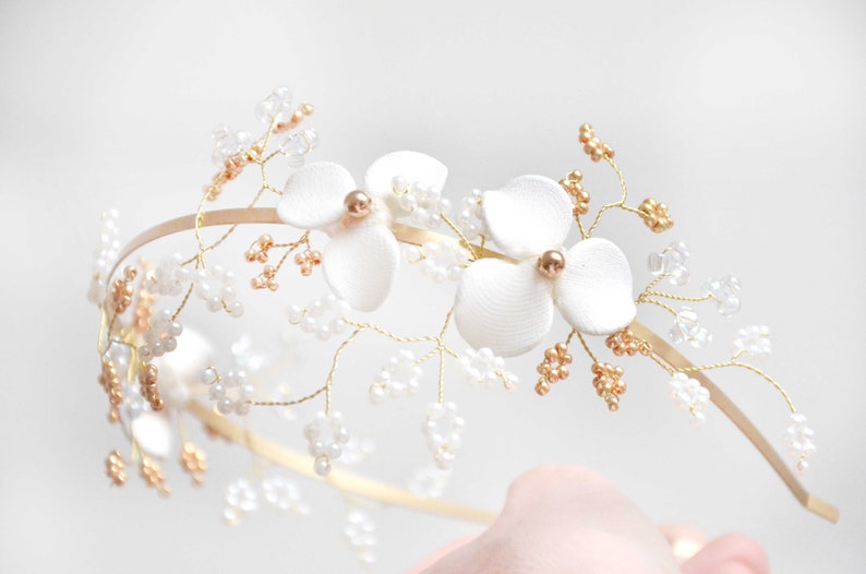 May include: A gold-toned metal headband featuring two white fabric flowers with gold centers, accented by delicate gold wire branches adorned with small clear beads and tiny gold-colored beads, creating a botanical-inspired hair accessory.