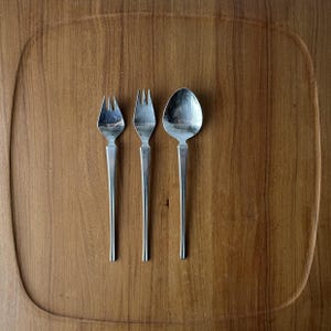 May include: A set of three stainless steel utensils consisting of two small forks with three tines each and one oval-shaped spoon. Each piece features a long, slender, flat handle that tapers toward the base, creating a uniform and sleek appearance.