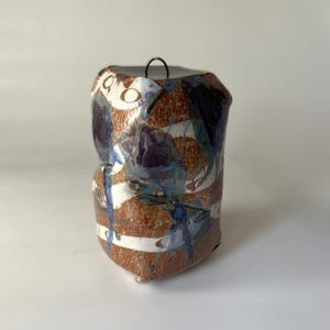 May include: A cylindrical ceramic vessel with a textured, mottled surface featuring a color palette of earthy brown, deep blue, and white. The piece has an irregular, hand-formed shape and includes a small, thin black metal loop attached to the top.