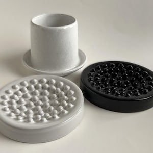May include: Three ceramic bathroom accessories: a white tumbler with a small base, and two soap dishes. One soap dish is white, the other is black. Both soap dishes have a textured, rounded surface.