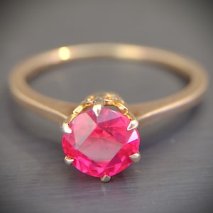 May include: A gold ring featuring a vibrant, cushion-cut, hot pink gemstone. The ring's band is a simple, polished gold, and the gemstone is held in place by delicate prongs. The ring is set against a neutral background.