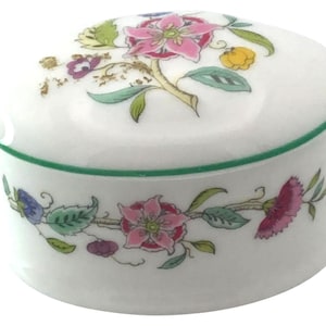 May include: A white, circular trinket box with a lid, decorated with a floral pattern in pink, green, yellow, and purple. The box has a green trim around the lid and base. The floral design includes detailed leaves and flowers.