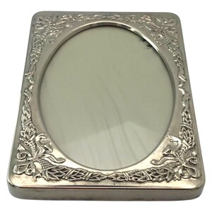 May include: Silver rectangular picture frame with an oval aperture. The frame has decorative floral and ribbon patterns around the edges. The frame is designed to hold a photograph or artwork. The frame is made of metal.