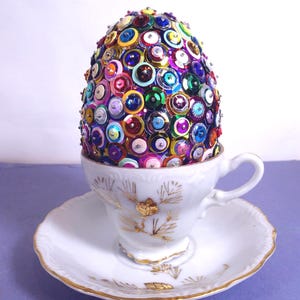 May include: A decorative egg covered in colorful sequins sits in a white teacup and saucer. The egg is adorned with various shades of sequins, including red, blue, green, and pink. The teacup and saucer have gold trim.
