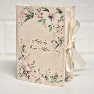 Op de afbeelding: Een klein, crèmekleurig doosje met een bloemenpatroon en de woorden "Happily Ever After" in gouden letters. Het doosje is versierd met een wit satijnen lint.