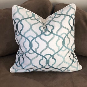 May include: White throw pillow with a teal and blue geometric pattern. The pillow has a white border and is made of a soft, textured fabric.