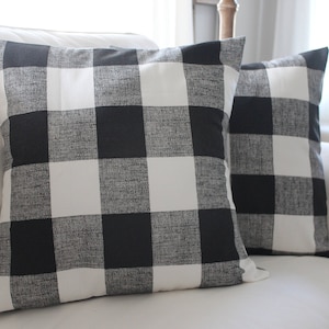May include: Two black and white buffalo check throw pillows on a white sofa. The pillows are made of a soft, textured fabric.