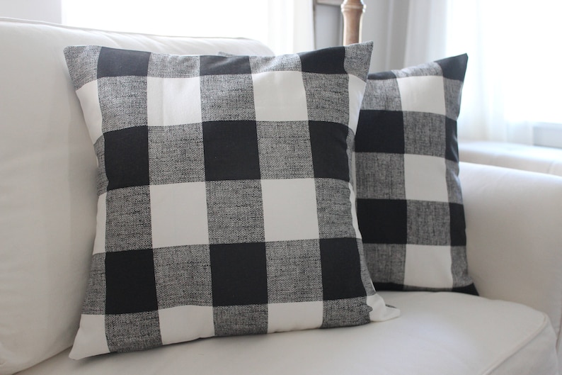May include: Two black and white buffalo check throw pillows on a white sofa. The pillows are made of a soft, textured fabric.