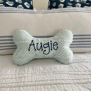 May include: A light blue and white dog bone shaped pillow with the name "Augie" embroidered in black thread. The pillow is on a white bed with a blue and white striped pillow behind it.