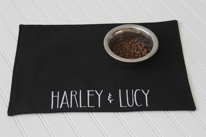 May include: Black pet food mat with white embroidered text "HARLEY & LUCY" and a stainless steel bowl filled with brown kibble.
