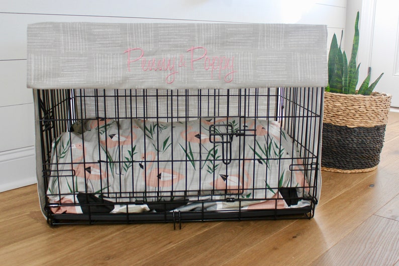 May include: A black metal dog crate with a gray fabric cover. The cover has the words "Penny & Poppy" embroidered on it in pink thread. The crate is lined with a white and pink patterned bedding.