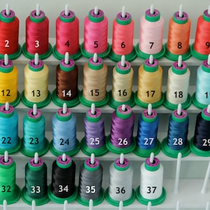 Puede incluir: Un conjunto de 37 carretes de hilo, organizados en filas en un estante blanco. Los carretes est&aacute;n numerados del 1 al 37. Los colores del hilo incluyen rojo, amarillo, azul, verde, morado, negro, blanco y beige.