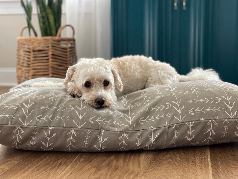 May include: A small white dog with brown eyes is lying on a grey dog bed with a white tree pattern. The dog bed is on a wooden floor.